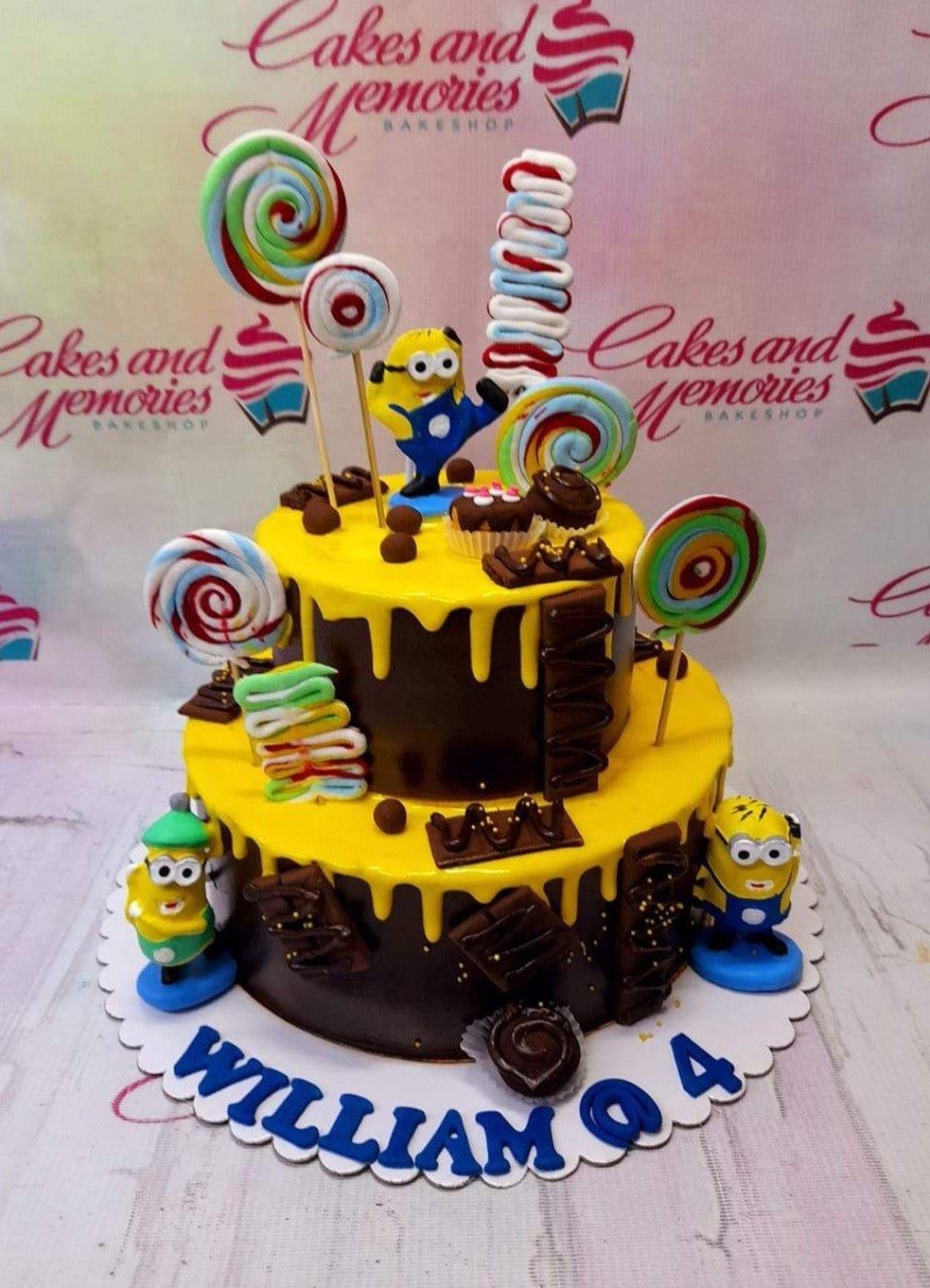 Two-tier Minions themed chocolate cake with yellow drip icing, lollipops, and chocolate bar decorations.