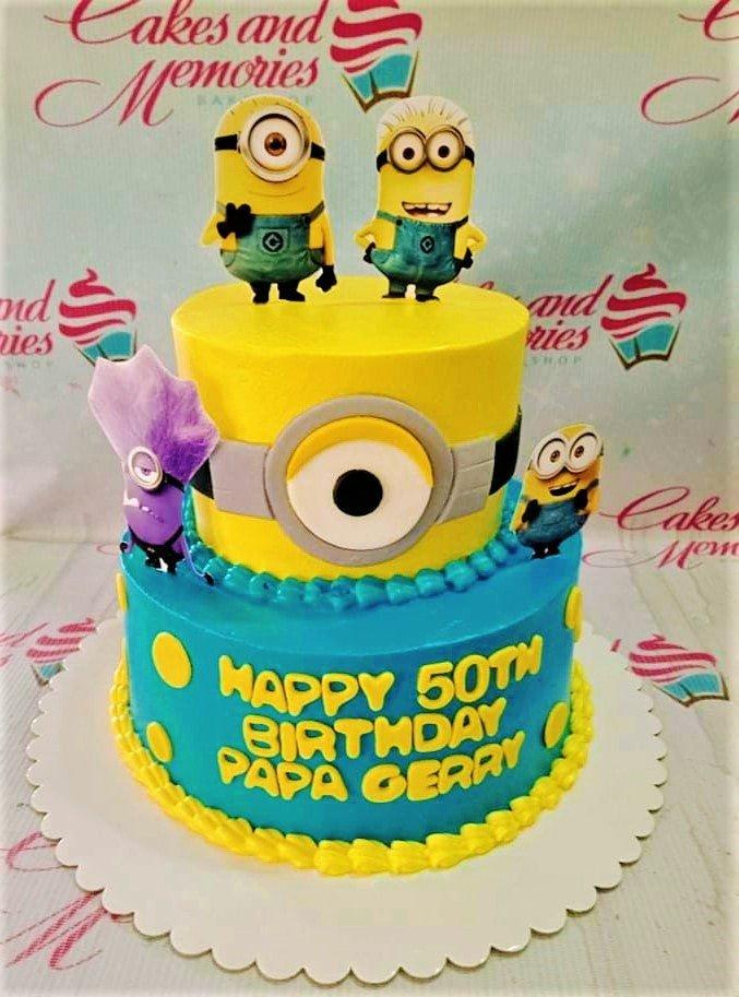 Two-tier Minions themed birthday cake with yellow and blue icing, featuring printed Minion characters and a large fondant eye design.