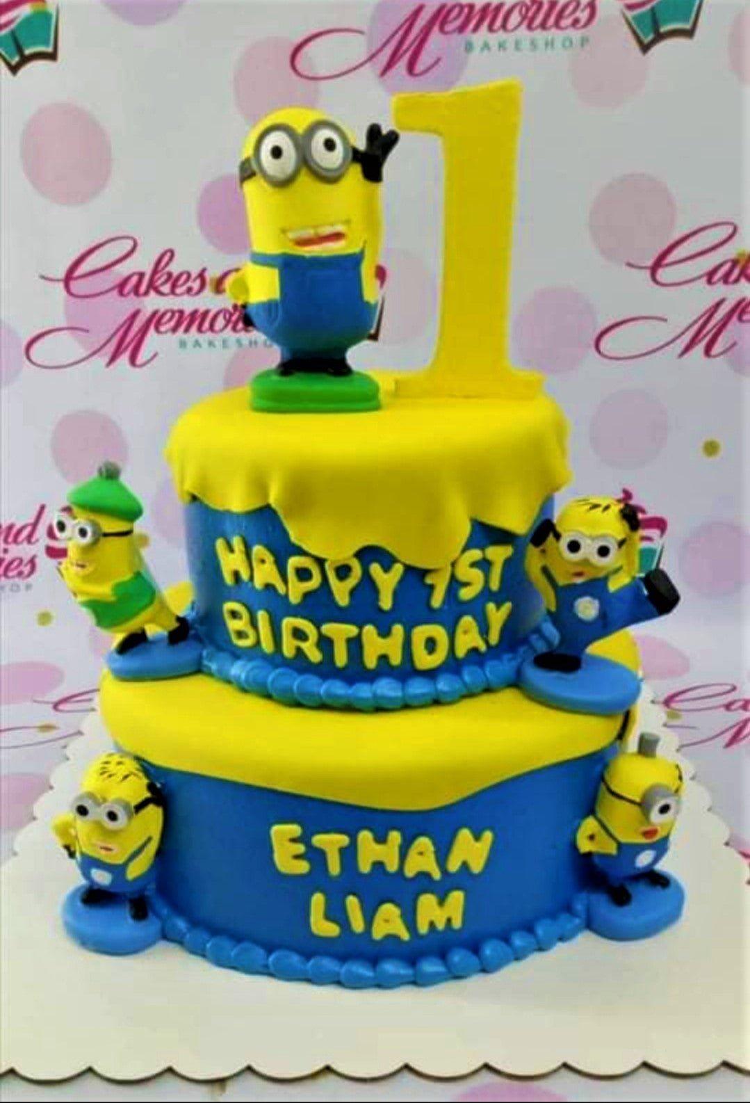 Two-tier blue and yellow Minions themed birthday cake with fondant figures and a large yellow number one topper.