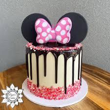 Minnie Mouse themed cake with white icing, chocolate drip, pink and white sprinkles, and a large Minnie Mouse bow topper with ears.