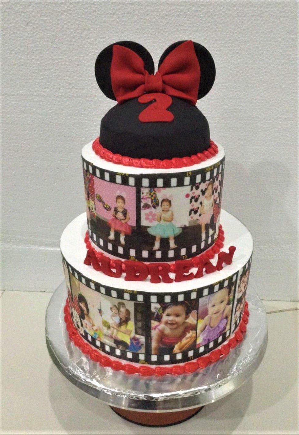 Two-tier Minnie Mouse themed birthday cake with film strip photo prints, red icing borders, and a black Minnie ear topper with a red bow.