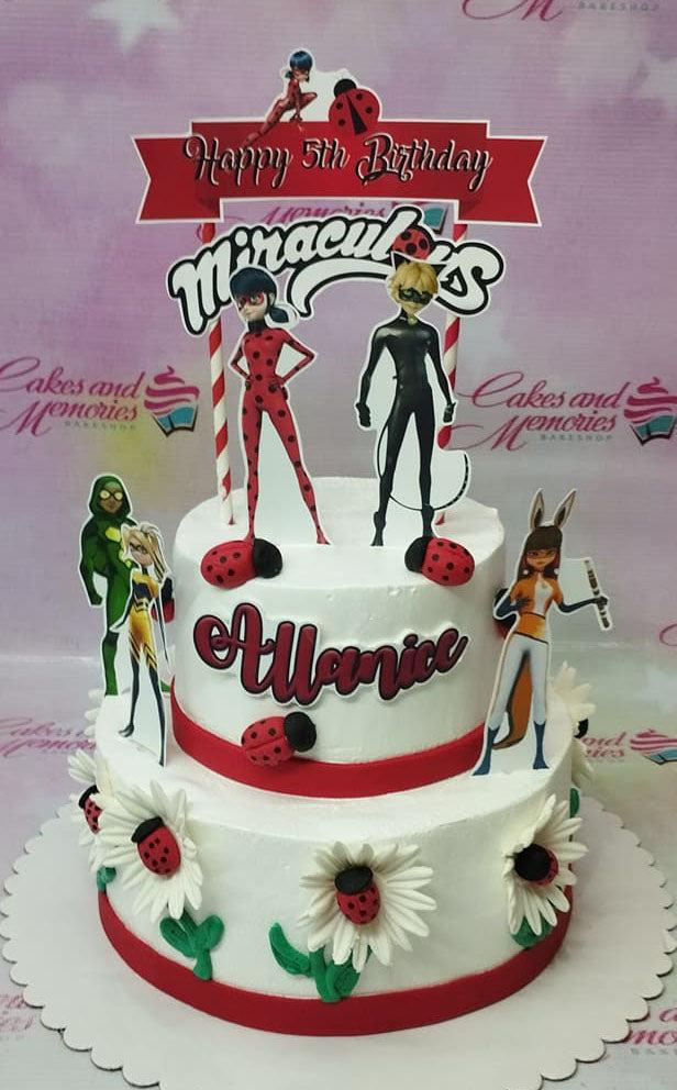 Two-tier white soft-icing Miraculous Ladybug themed birthday cake with character printouts, ladybug accents, and white daisies.