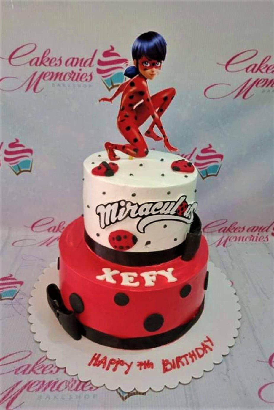 Two-tier Miraculous Ladybug themed cake with red and white icing, black polka dots, and a Ladybug character topper.