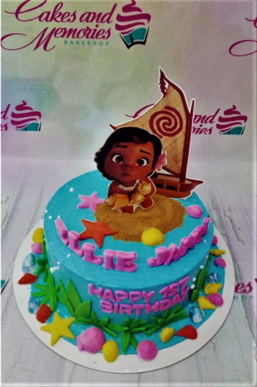 Moana themed birthday cake with a blue icing base, sand texture, and character printouts of Moana and her boat.