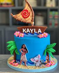 Moana themed blue cake with a large sailboat topper, palm trees, and Moana and Pua character cutouts.