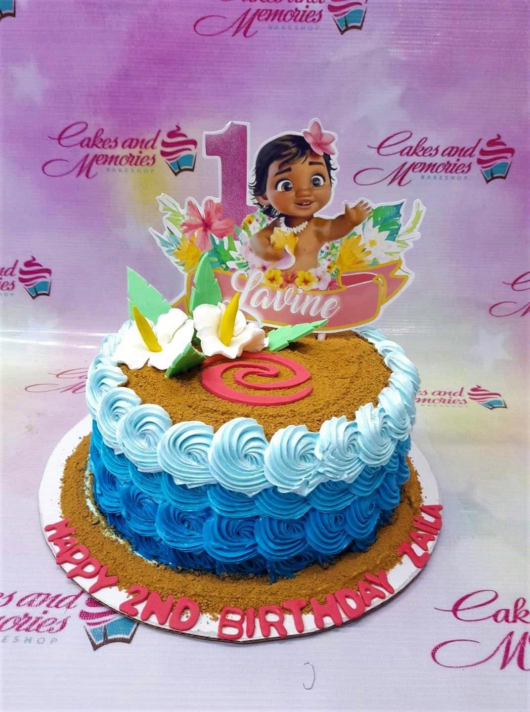 Moana themed birthday cake with blue wave icing, sand-like crumbs, and a Moana character printout topper.