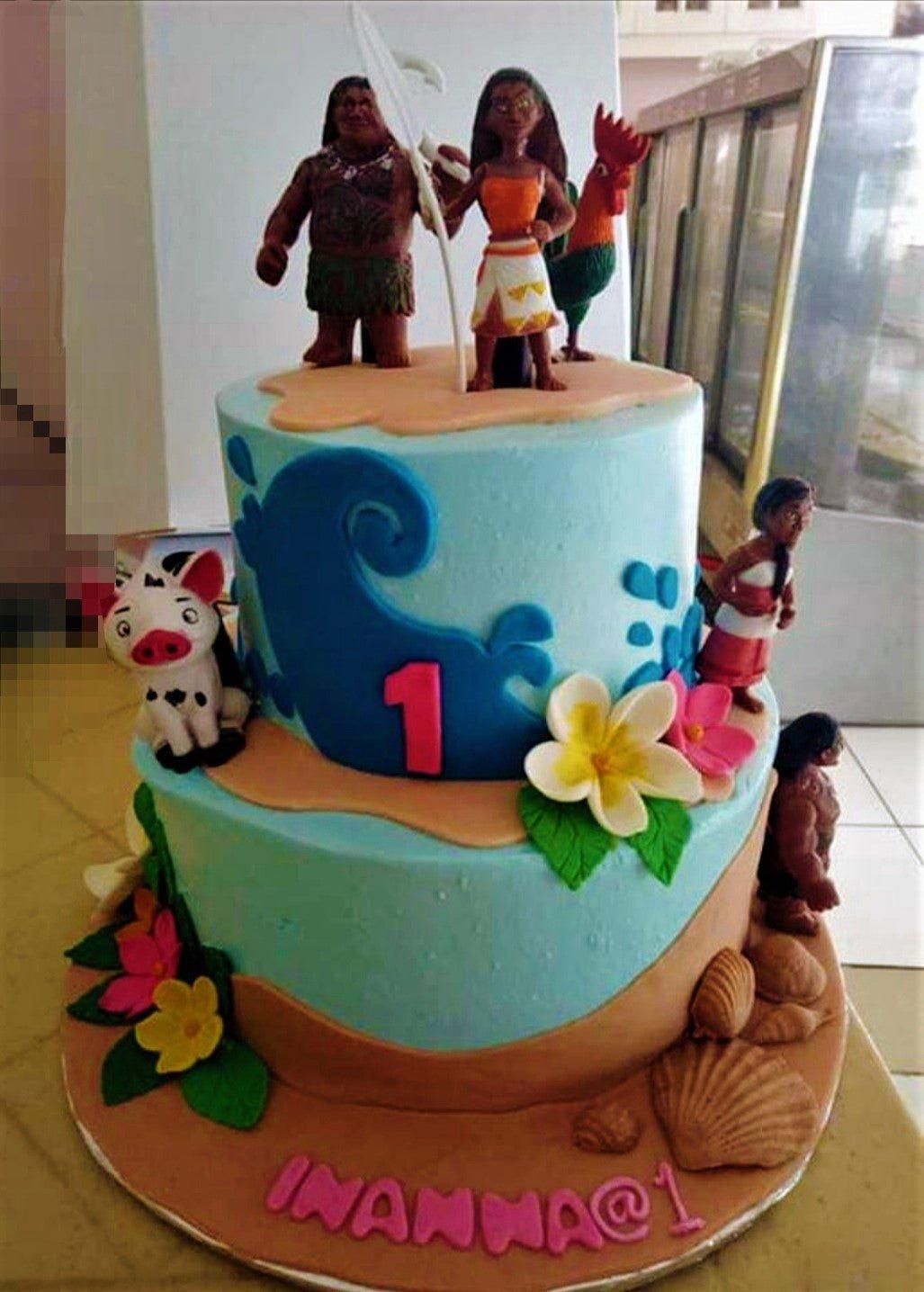 Two-tier Moana themed birthday cake with Maui, Moana, and Hei Hei figurines, fondant waves, and tropical flowers.