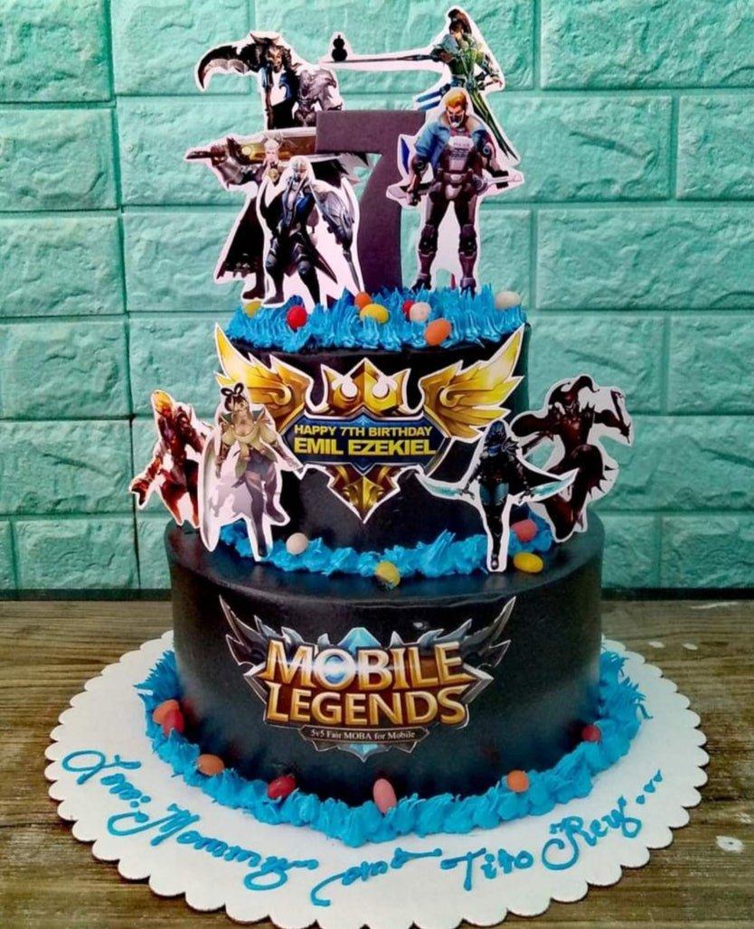 Two-tier black Mobile Legends themed cake with blue icing borders, character printouts, and a large purple number seven topper.