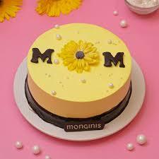 Yellow round cake for Mom featuring a large yellow icing sunflower, chocolate letters, and white pearl sprinkles on a pink background.