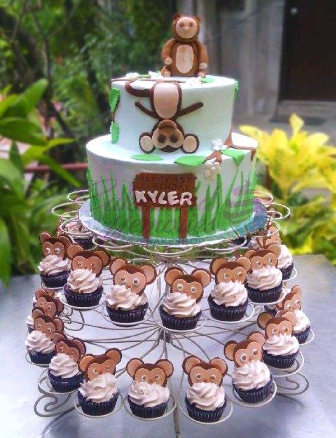 Two-tier light blue monkey themed birthday cake with fondant monkey figures, green grass icing, and matching monkey cupcakes.