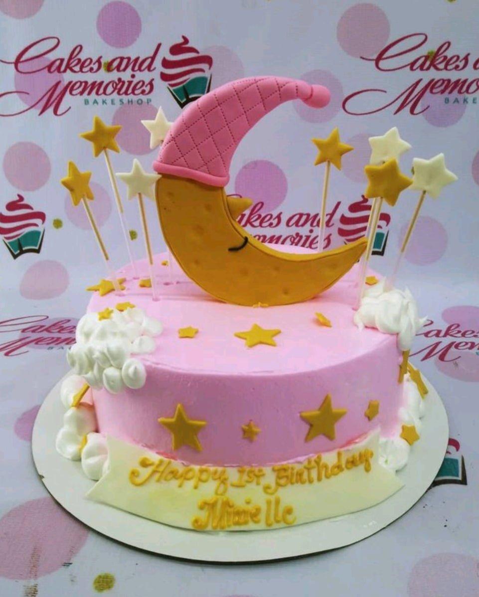 Pink 1st birthday cake with a large yellow crescent moon topper wearing a pink nightcap, surrounded by white and gold stars and fluffy white icing clouds.
