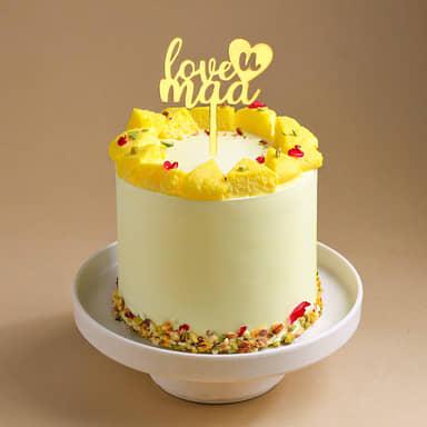 Tall light yellow cake topped with yellow sponge cake pieces, crushed pistachios, and a gold 'love u maa' topper.