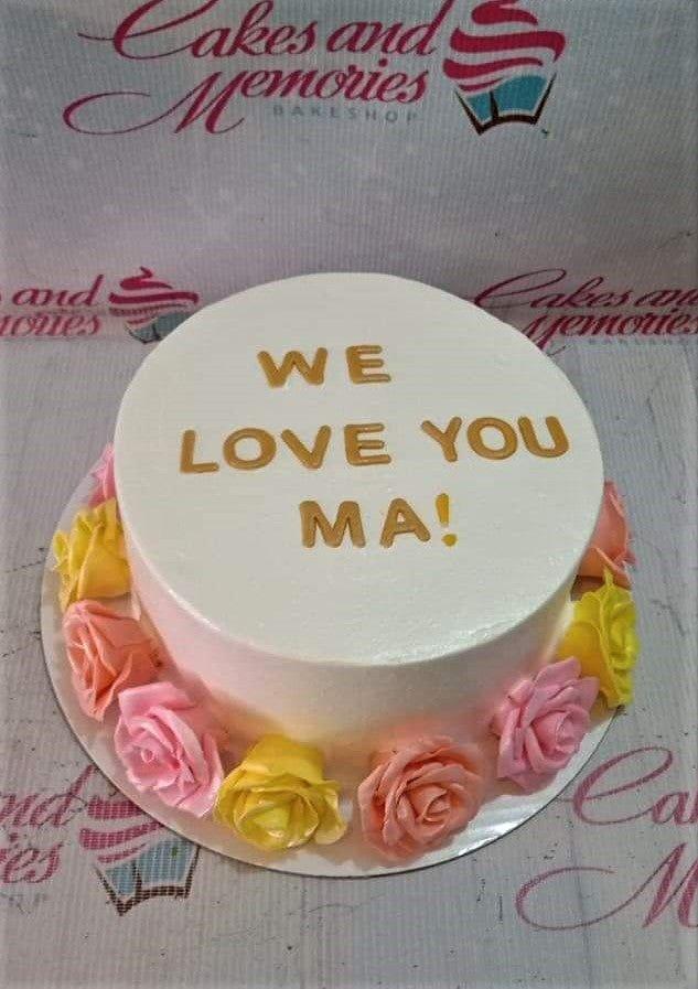 White minimalist cake with pink and yellow icing roses around the base and a gold message on top.