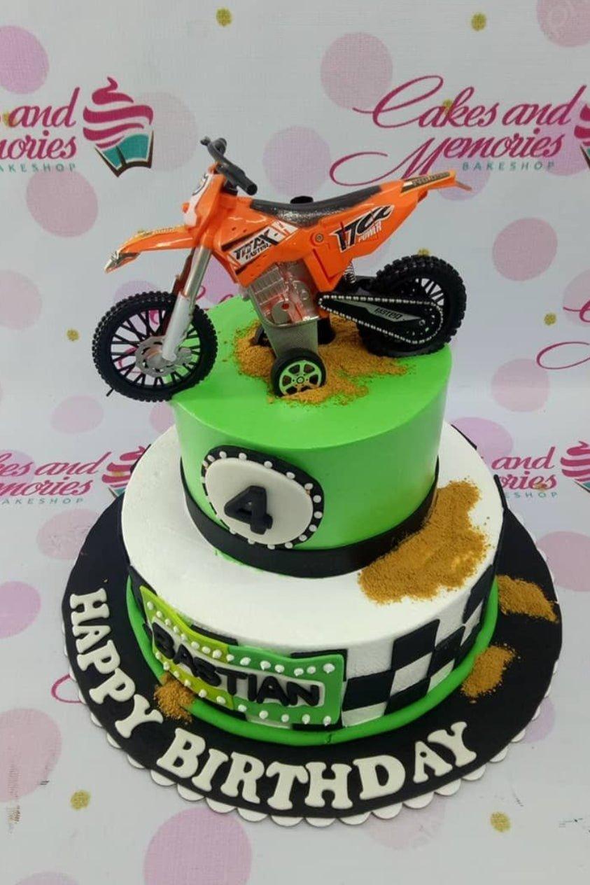 Two-tier motocross birthday cake with a green top tier, white bottom tier, orange toy motorcycle topper, and checkered flag patterns.
