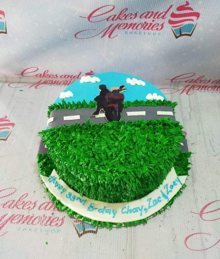 A round birthday cake featuring a scenic road design with a motorcycle printout topper and green grass-textured icing.