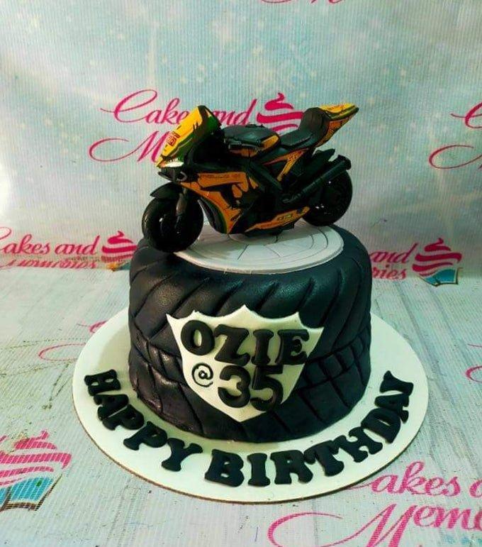 Black tire-shaped fondant cake with a detailed yellow and black motorcycle topper and a white shield message plaque.