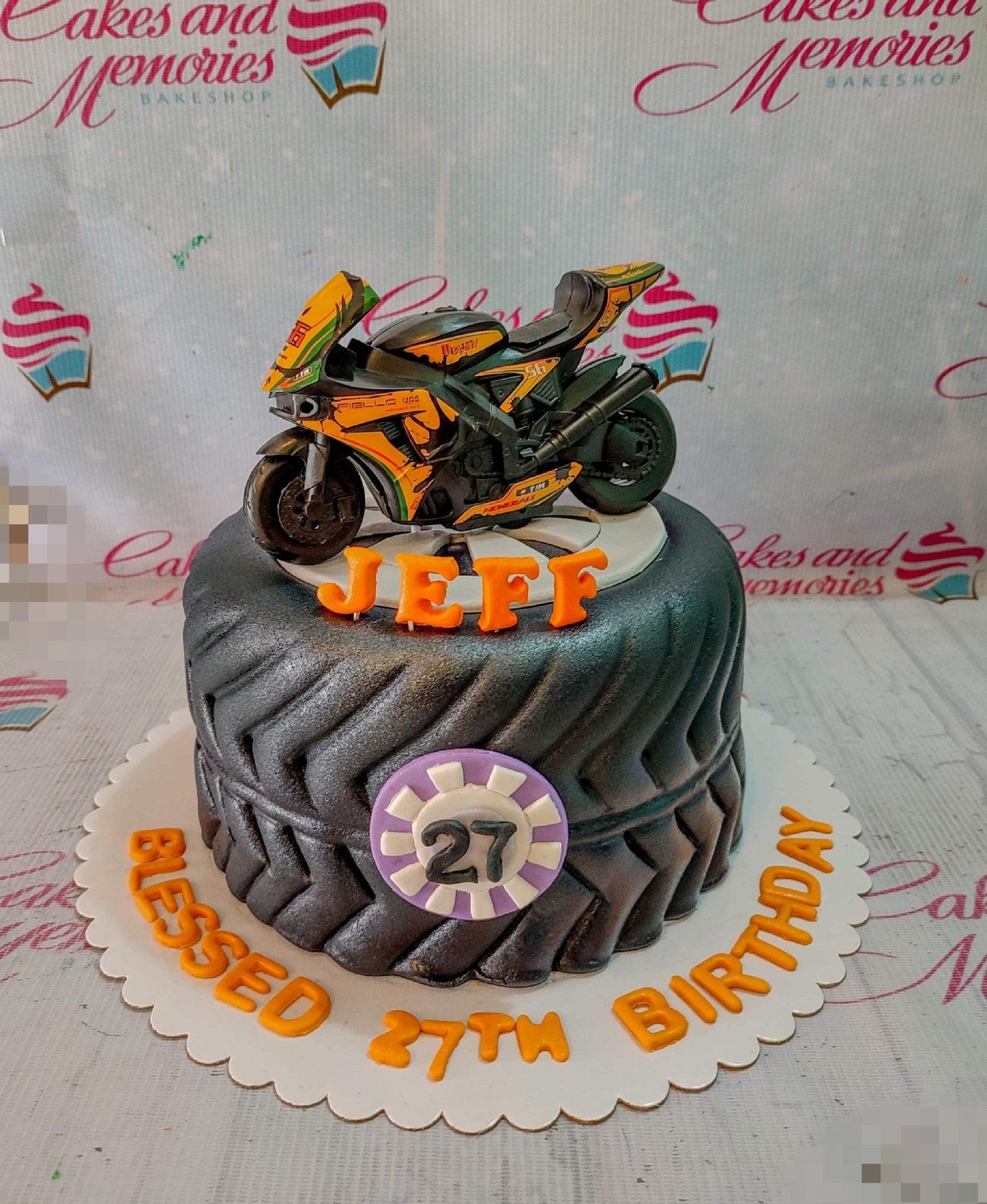 Black tire-shaped fondant cake with a detailed motorcycle toy topper and orange gumpaste letters for a 27th birthday celebration.