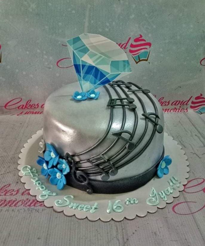 Silver metallic fondant cake with black musical notes and blue flowers, topped with a large blue diamond printout.