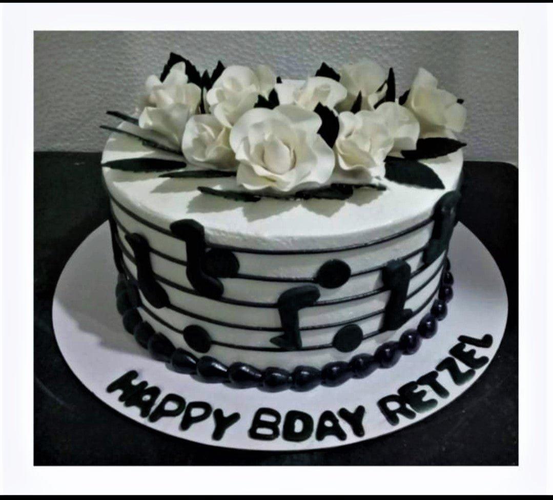 White round cake with music note designs on the side and white gumpaste roses on top, featuring a black birthday message on the base board.