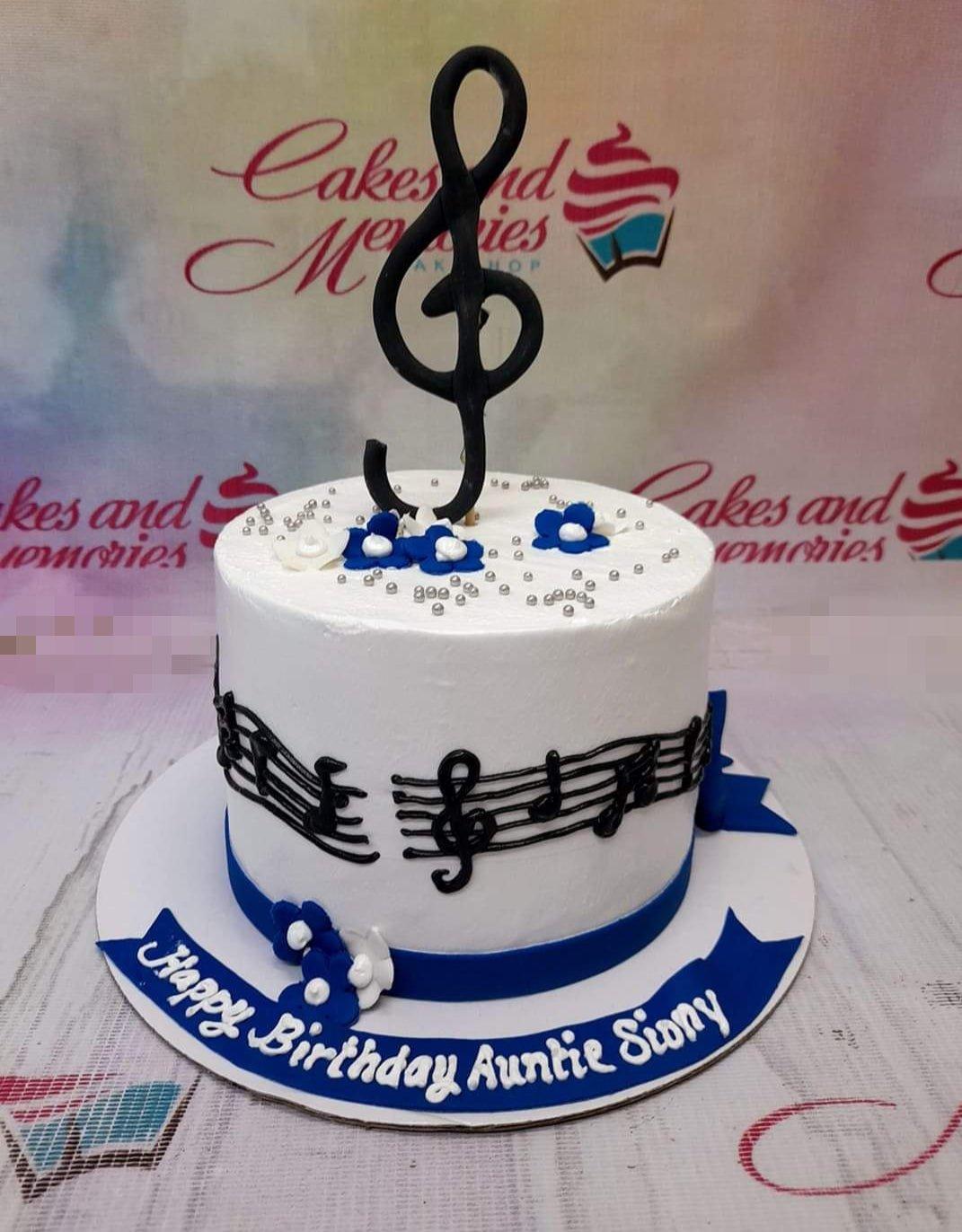 White 1-tier music themed birthday cake with black musical notes, blue ribbon, and a large black treble clef topper.