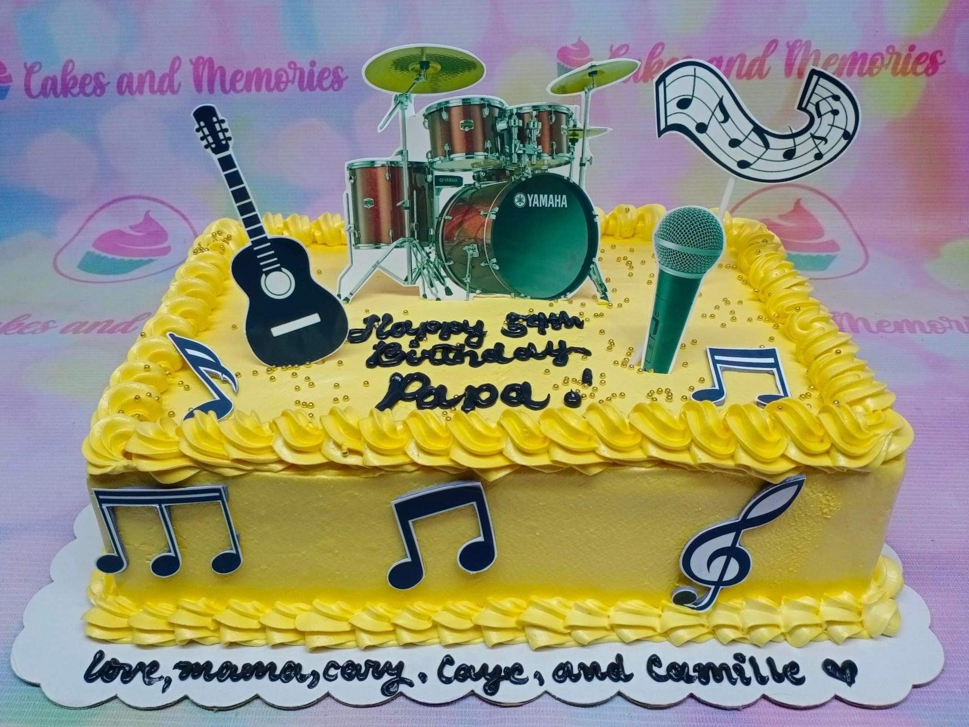 Yellow rectangular music themed cake with drum set, guitar, and microphone toppers, featuring black icing text and music note decorations.