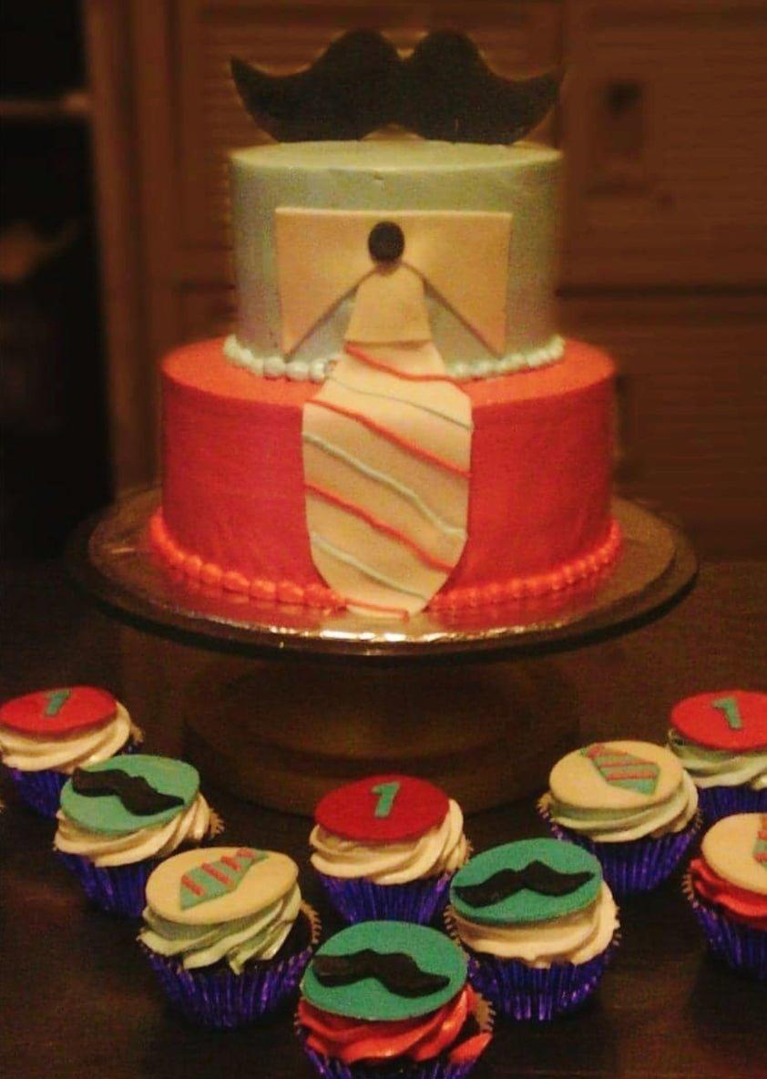Two-tier tuxedo and mustache themed cake with a light blue top tier and red bottom tier, featuring a fondant tie and mustache topper.