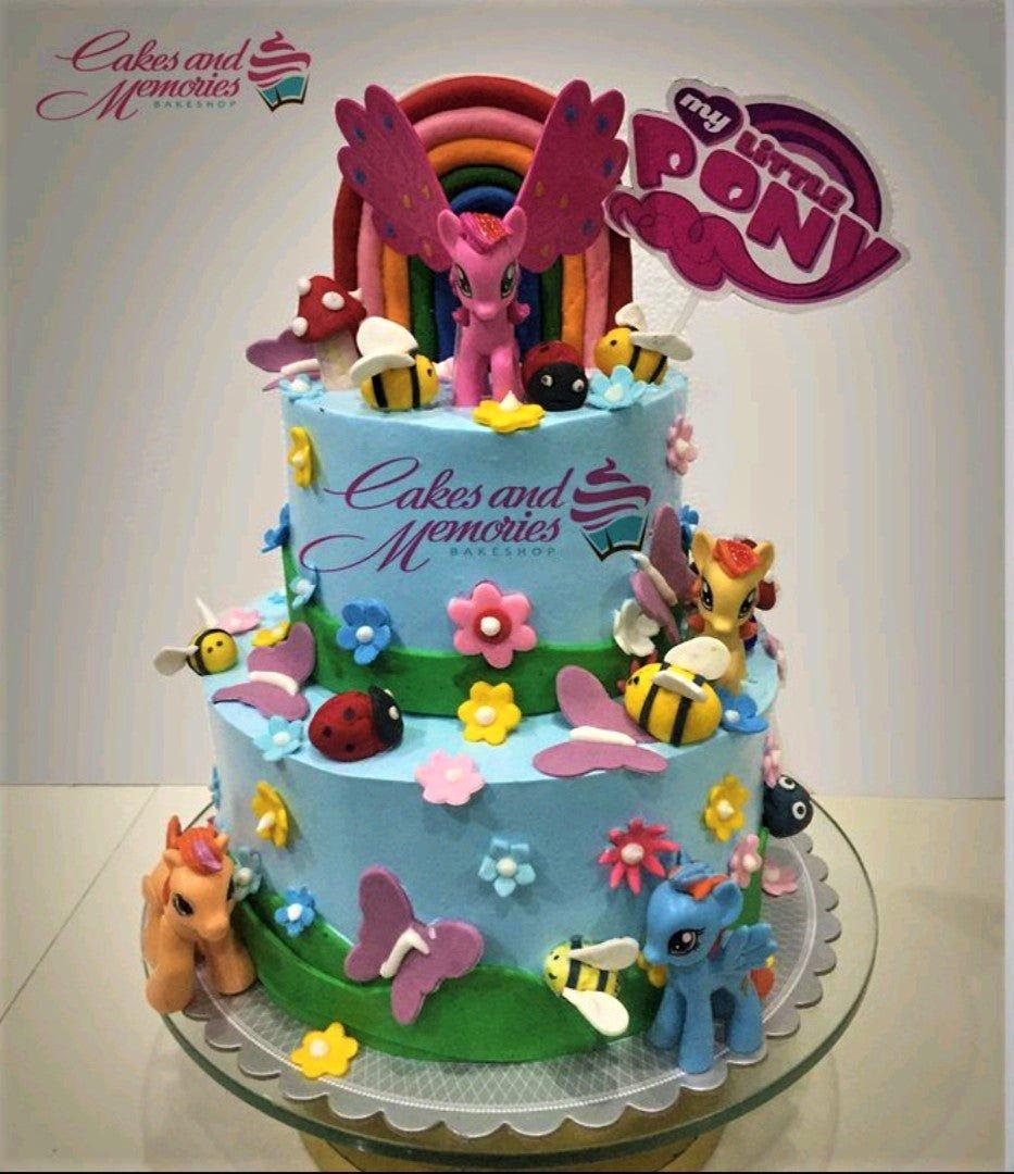 Two-tier light blue My Little Pony themed cake with fondant rainbow, pony figures, bees, ladybugs, and flowers.