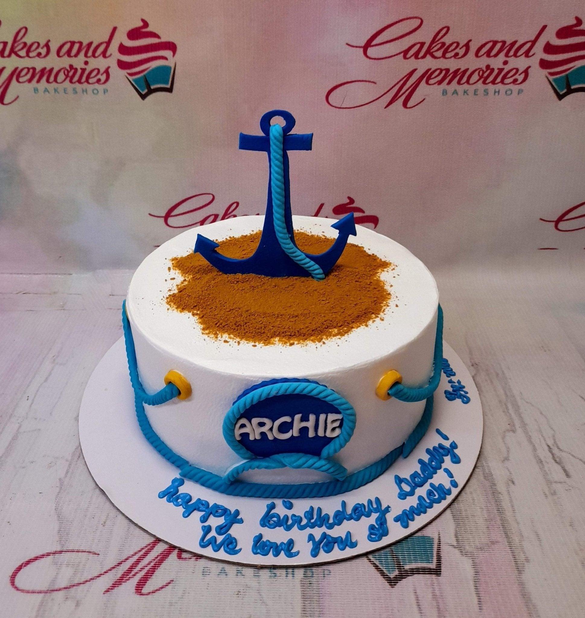 Nautical themed birthday cake with a large blue anchor topper on a sand-like surface and blue rope icing details.