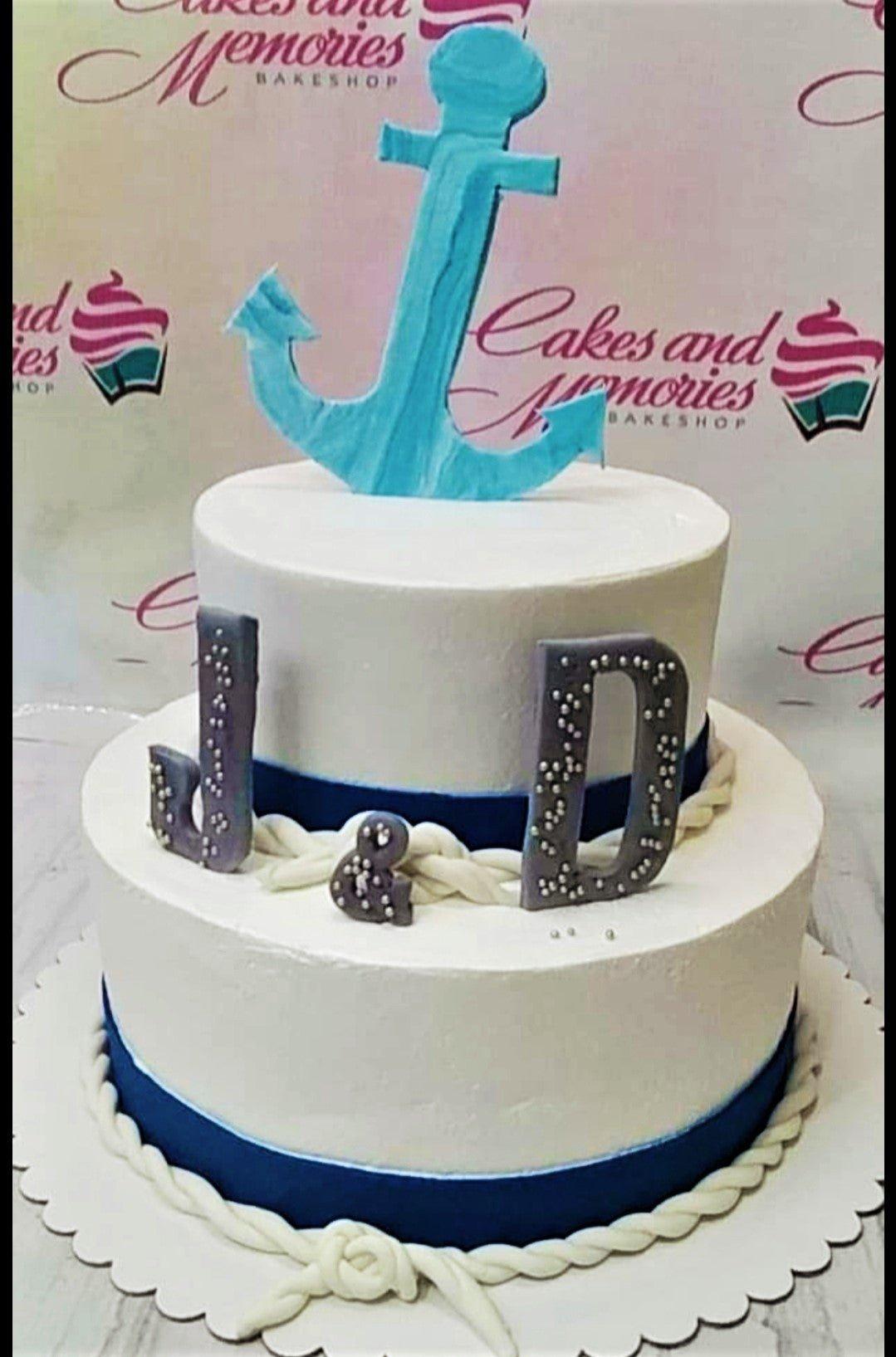 Two-tier white nautical themed cake with a large light blue anchor topper and grey initials J and D with silver pearls.
