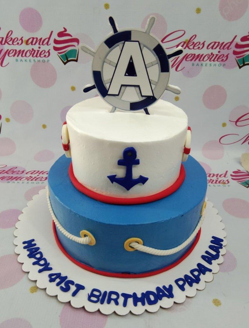 Two-tier nautical themed birthday cake in white and blue with anchor and ship wheel toppers.