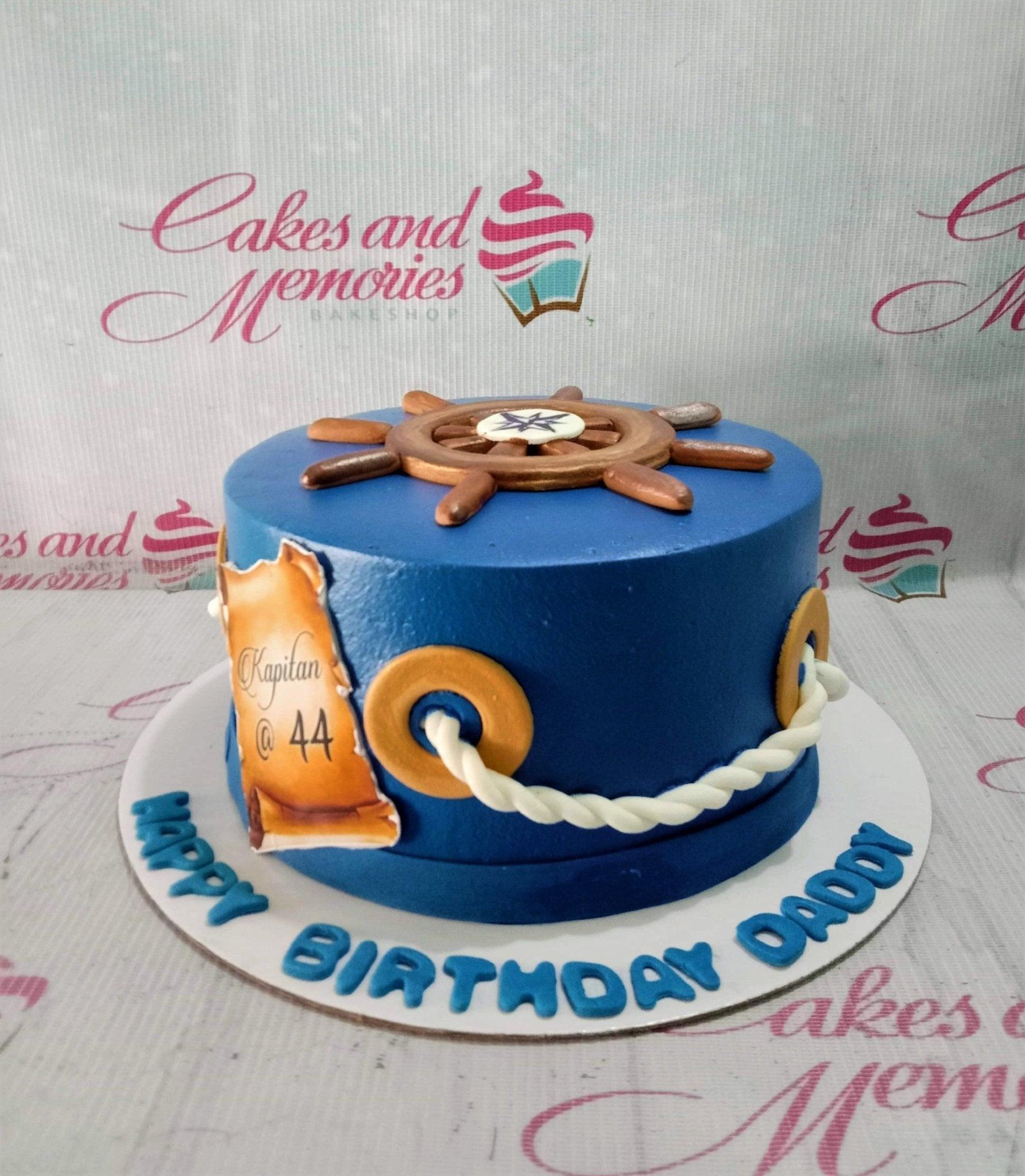 Blue nautical themed birthday cake for a captain featuring a ship wheel topper, rope details, and a scroll message.
