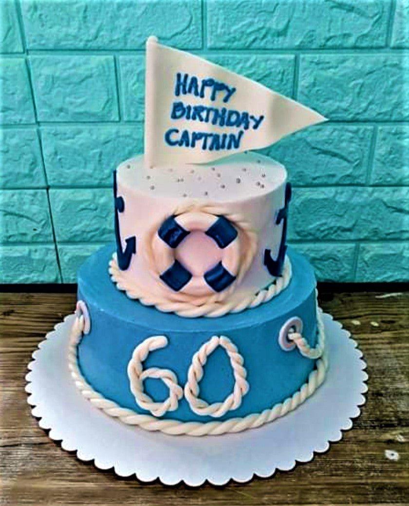 Two-tier nautical themed birthday cake in light blue and white with fondant rope, anchor, and lifebuoy decorations.