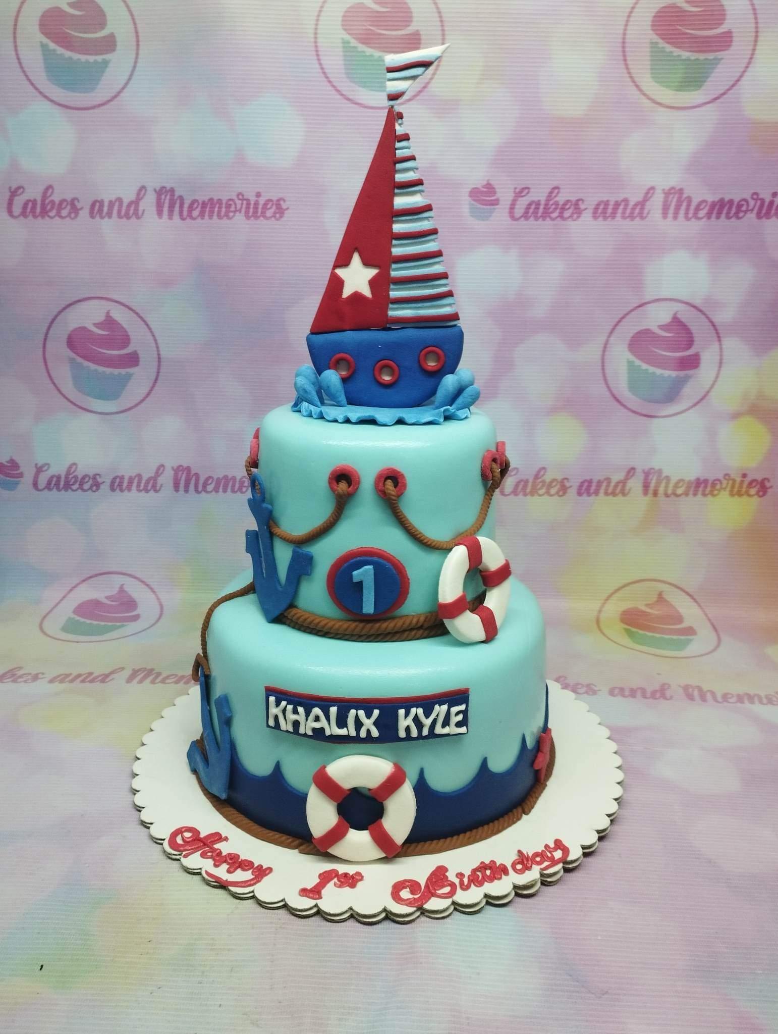 Two-tier nautical themed birthday cake in light blue fondant with a red and blue sailboat topper, anchors, and lifebuoys.