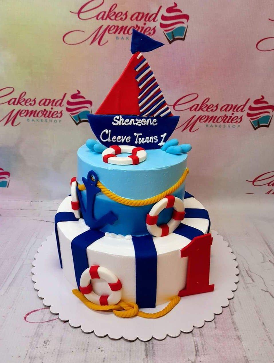 Two-tier nautical themed birthday cake with a red and blue sailboat topper, anchors, and lifebuoys on blue and white striped icing.