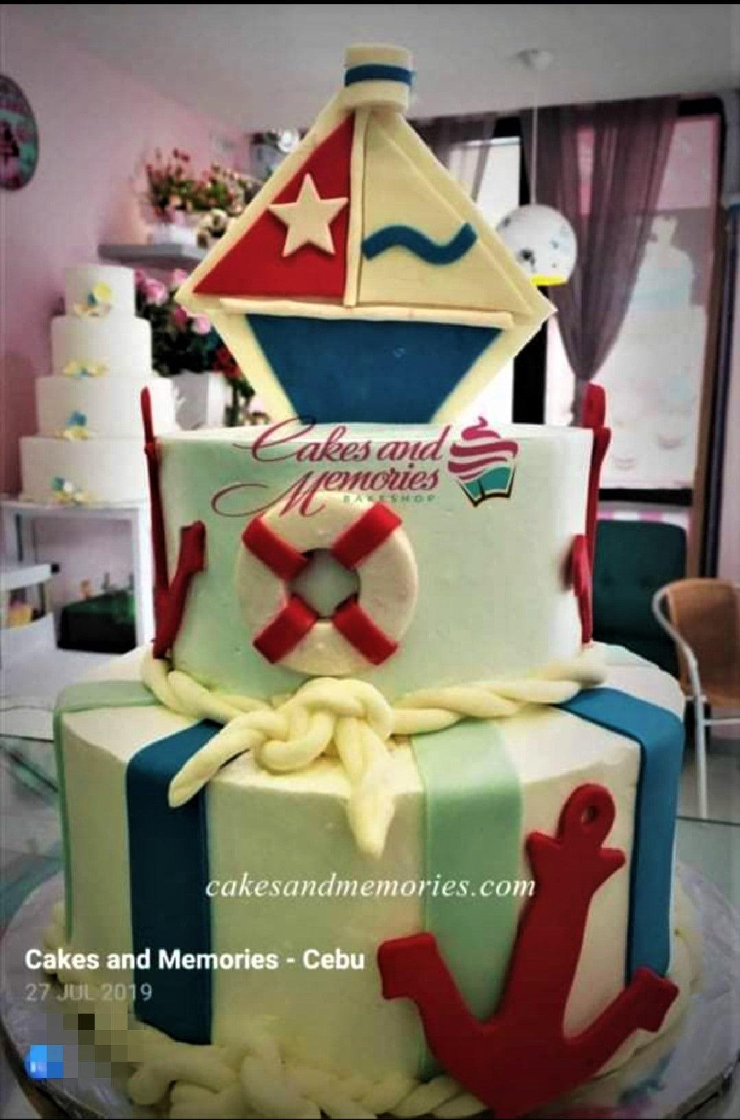 Nautical themed 2-tier fondant cake with a sailboat topper, lifebuoy, anchor, and rope details in red, blue, and mint green.