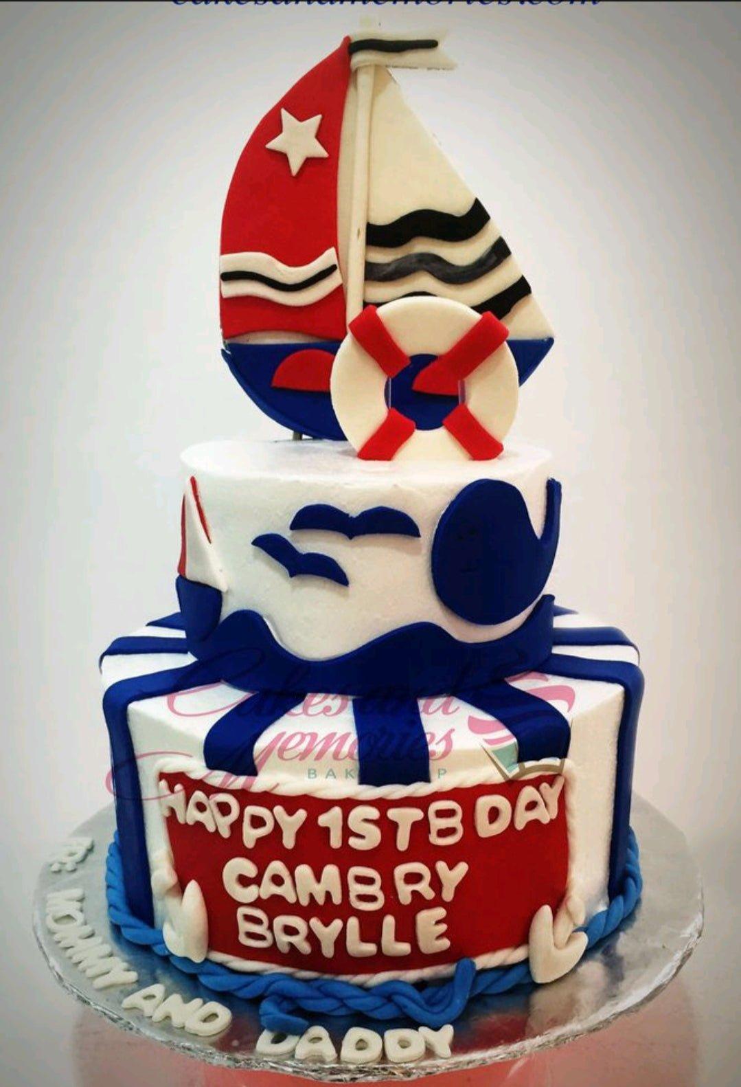 Two-tier nautical themed birthday cake in red, white, and blue with a large fondant sailboat topper and whale decorations.