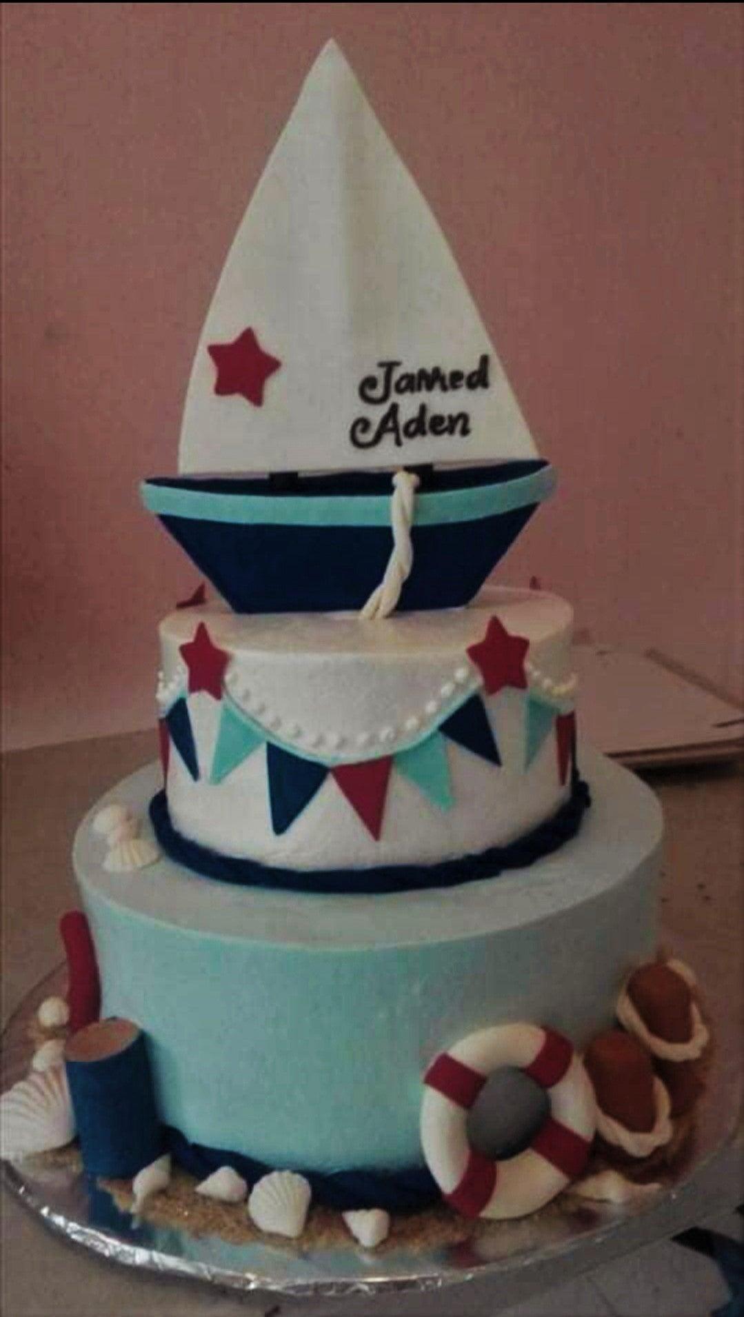 Two-tier nautical sailboat cake in light blue and white with fondant sailboat topper, lifebuoy, and seashell decorations.