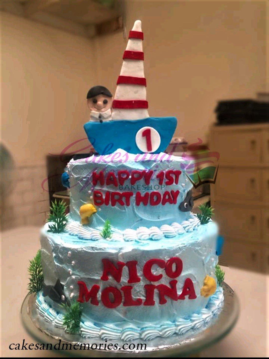 Two-tier light blue nautical themed cake with a fondant boat topper, sailor figure, and red gumpaste lettering.