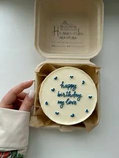 White bento cake with navy blue icing text and tiny heart decorations in a compostable box.