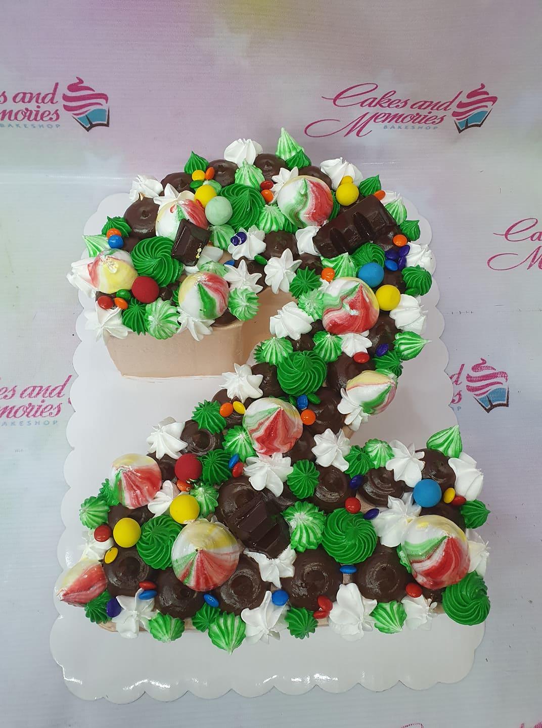 Number 2 shaped cake decorated with green and white icing rosettes, chocolate pieces, and colorful candy toppings.