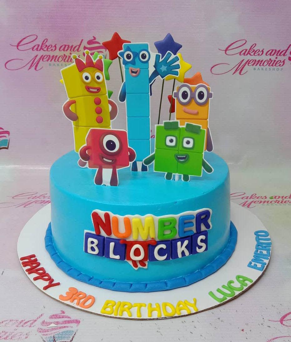 Light blue Numberblocks themed birthday cake with colorful character printouts and gumpaste letters on a white base board.