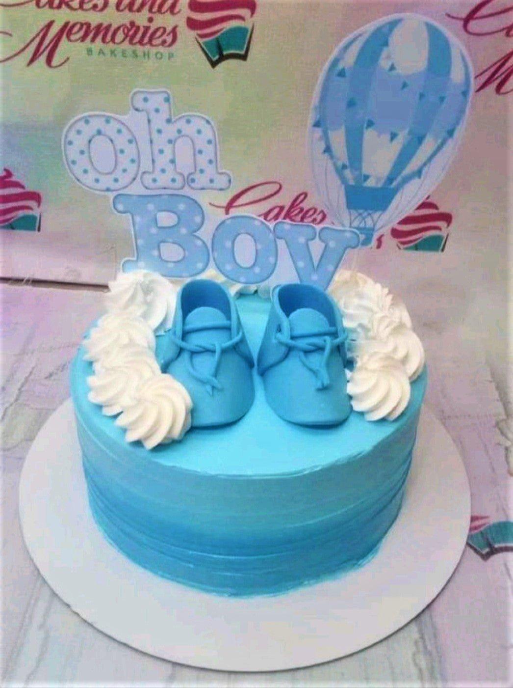 Light blue 1-tier baby boy cake with fondant baby shoes, white icing swirls, and a hot air balloon printout topper.