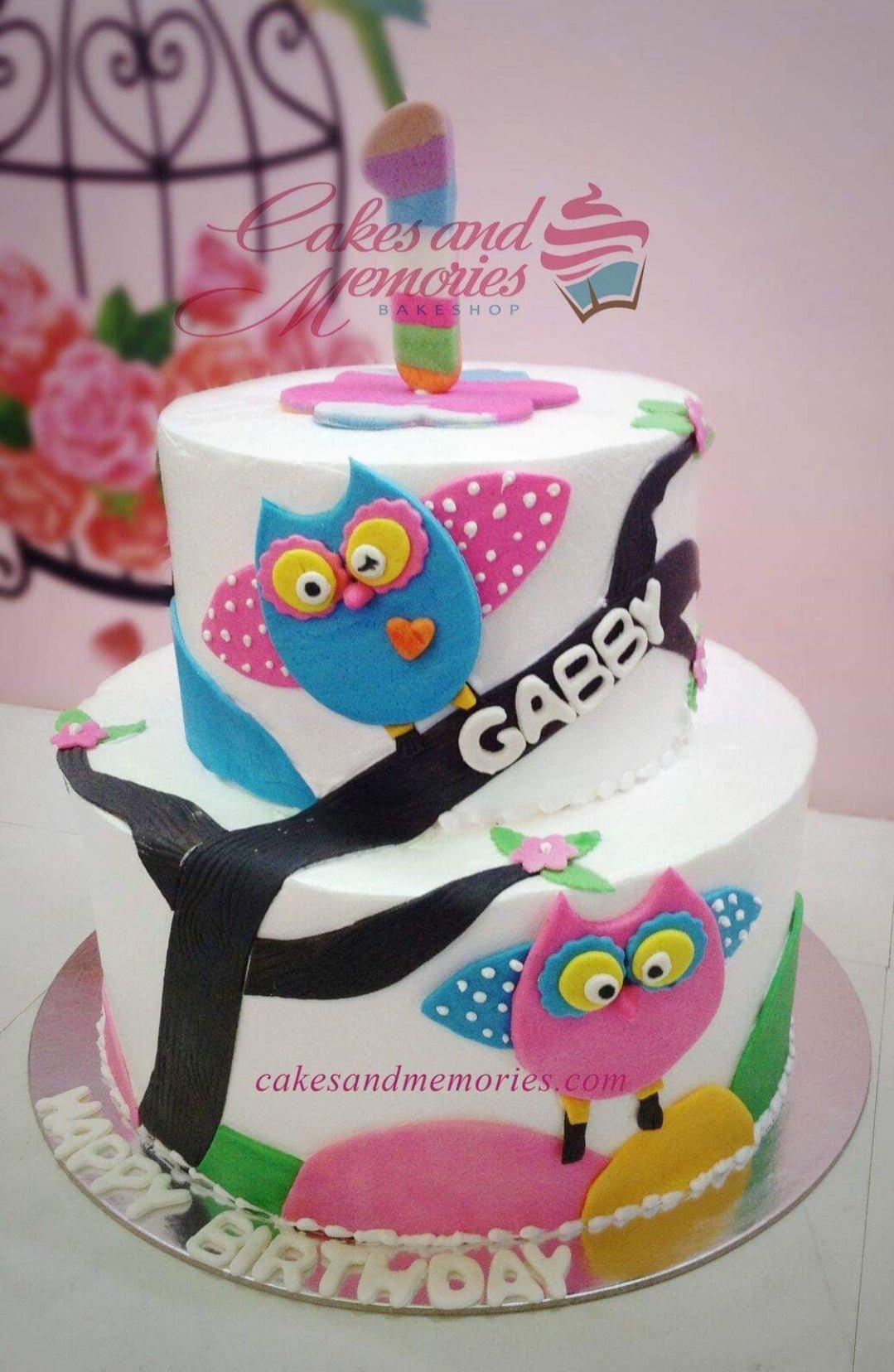 Two-tier white owl themed birthday cake with colorful fondant owls on branches and a rainbow number one topper.