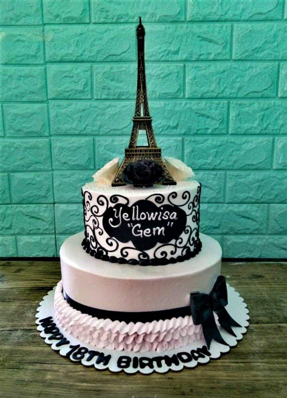Elegant 2-tier Paris themed birthday cake with a black Eiffel Tower topper, white icing, and black scrollwork designs.
