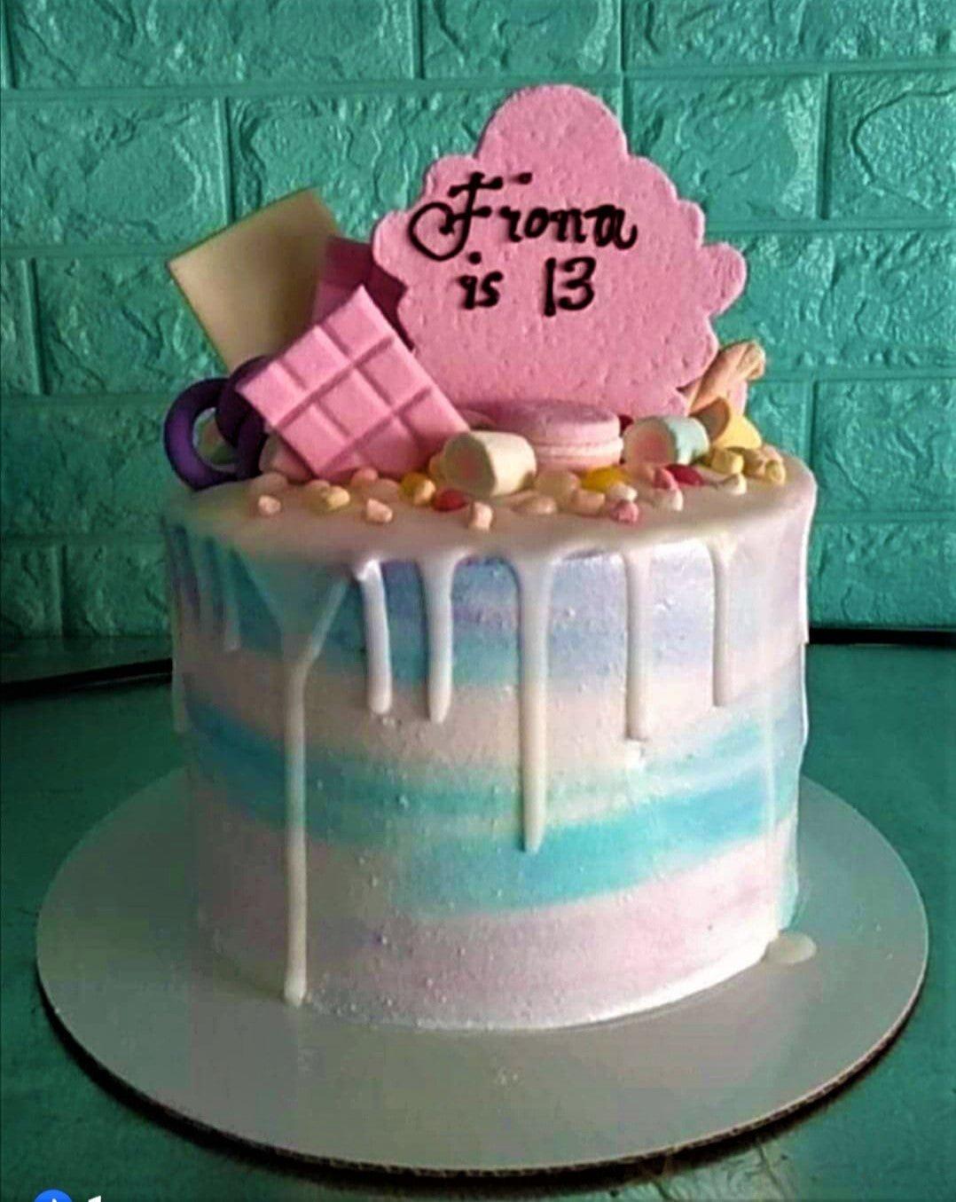 A single tier birthday cake with pastel blue and pink gradient icing, white chocolate drip, and various candy toppers including a pink name plaque.