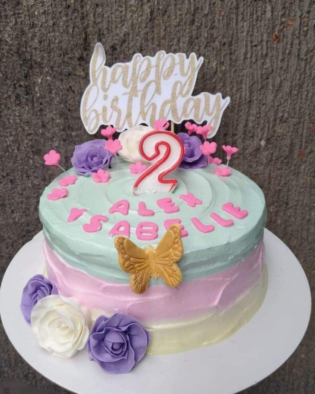 A pastel tricolor birthday cake with mint, pink, and yellow icing, topped with purple and white roses, a gold butterfly, and a number 2 candle.