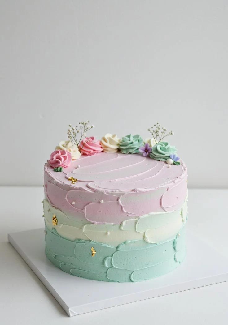 A pastel 1-tier cake with pink, mint, and white textured icing, topped with colorful icing rosettes, small purple flowers, and gold leaf accents.