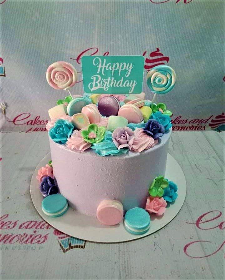 Lavender birthday cake decorated with pastel macarons, marshmallows, and colorful icing flowers with a teal Happy Birthday topper.