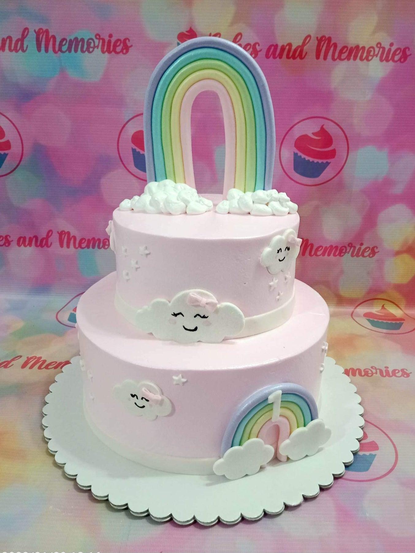 Two-tier light pink cake with a large pastel rainbow topper, fondant clouds with faces, and a small rainbow with the number 1.