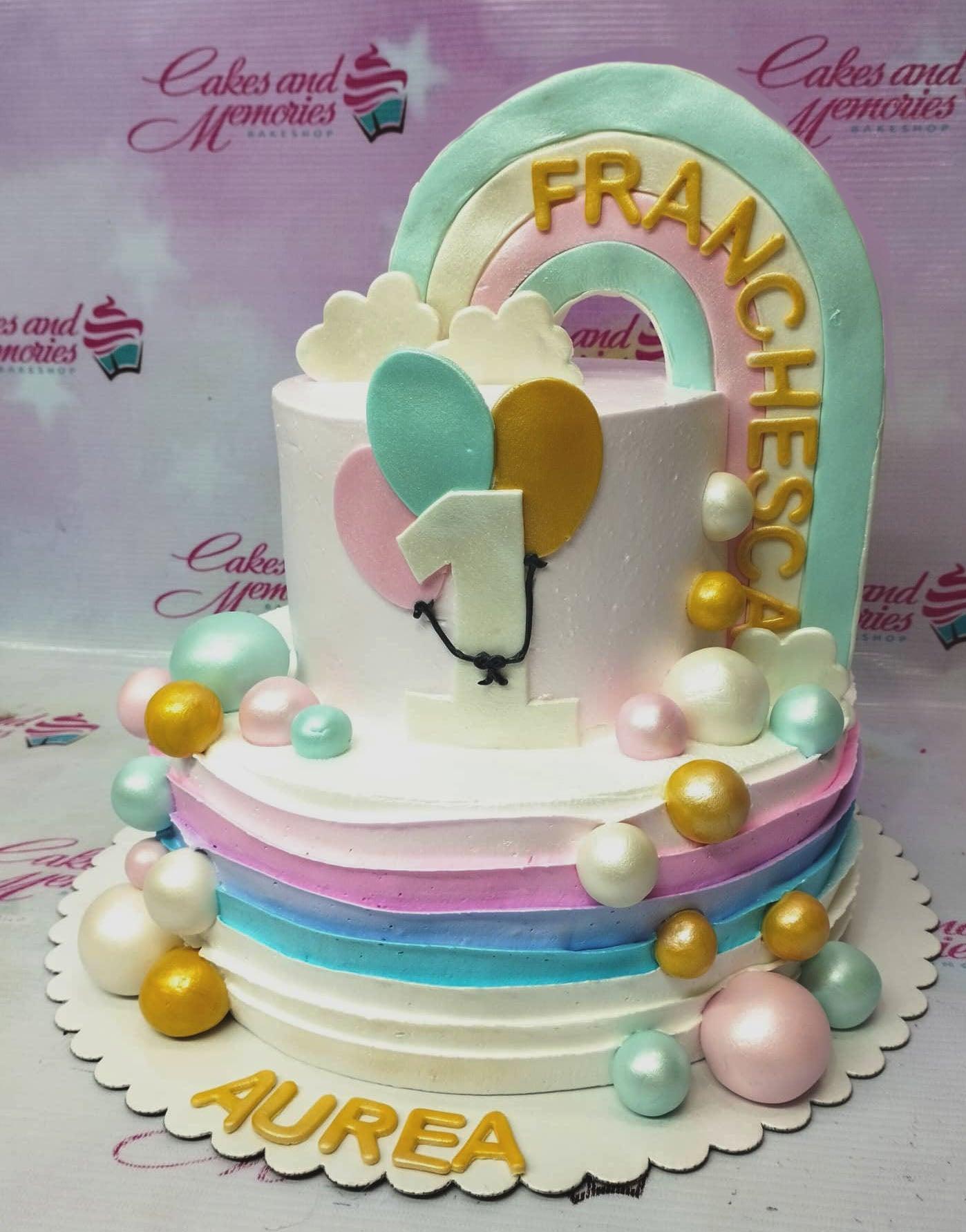 Two-tier pastel rainbow birthday cake with a large fondant rainbow topper, balloons, and colorful spheres.